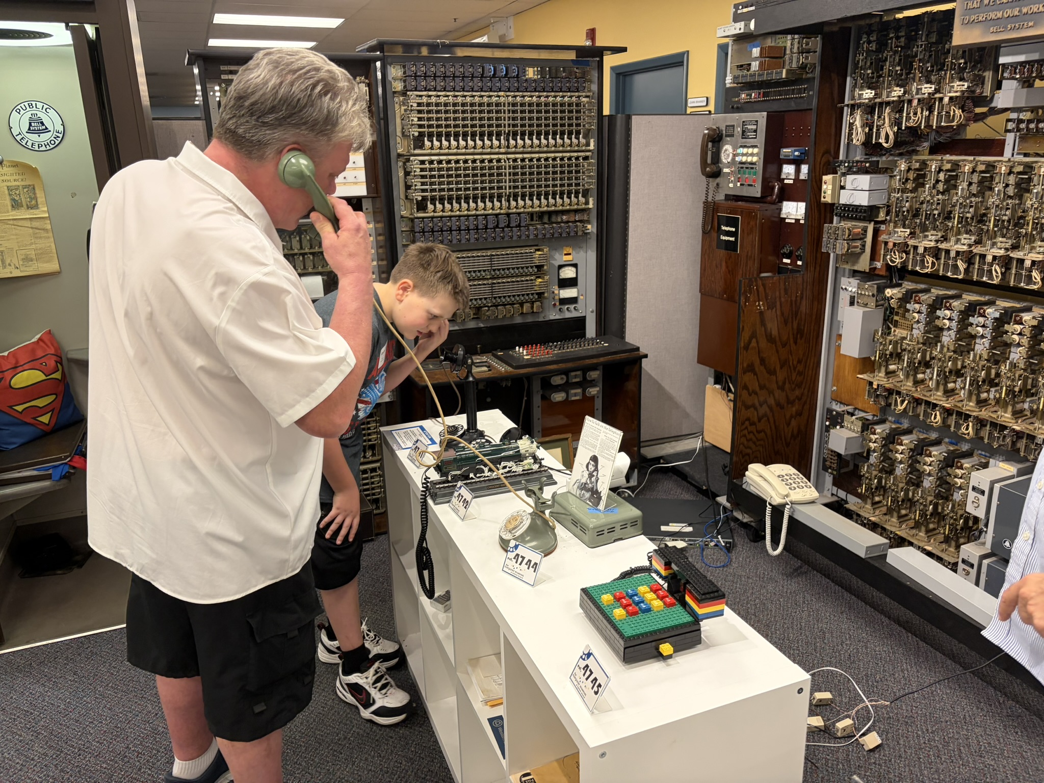 a man and boy talking on telephones in front of a big telephone relay system